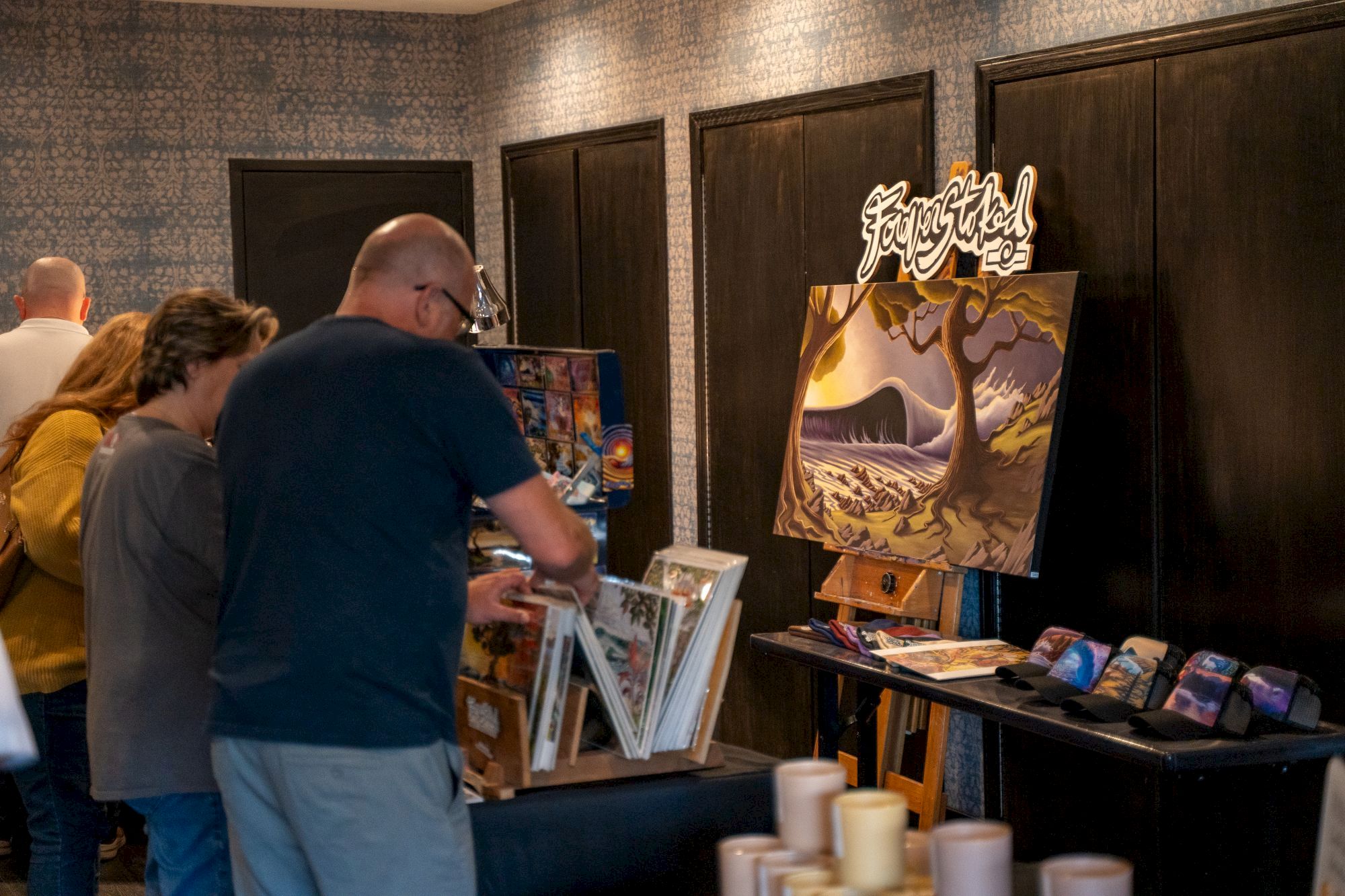 A group painting session at an art convention, with an easel and canvases showing a fantasy forest scene in the room.