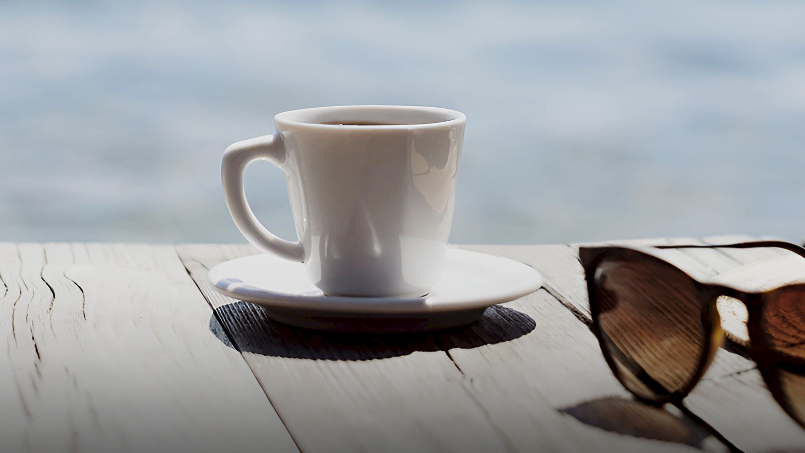A white coffee cup on a saucer sits on a weathered table beside a straw hat in the seaside breeze.
