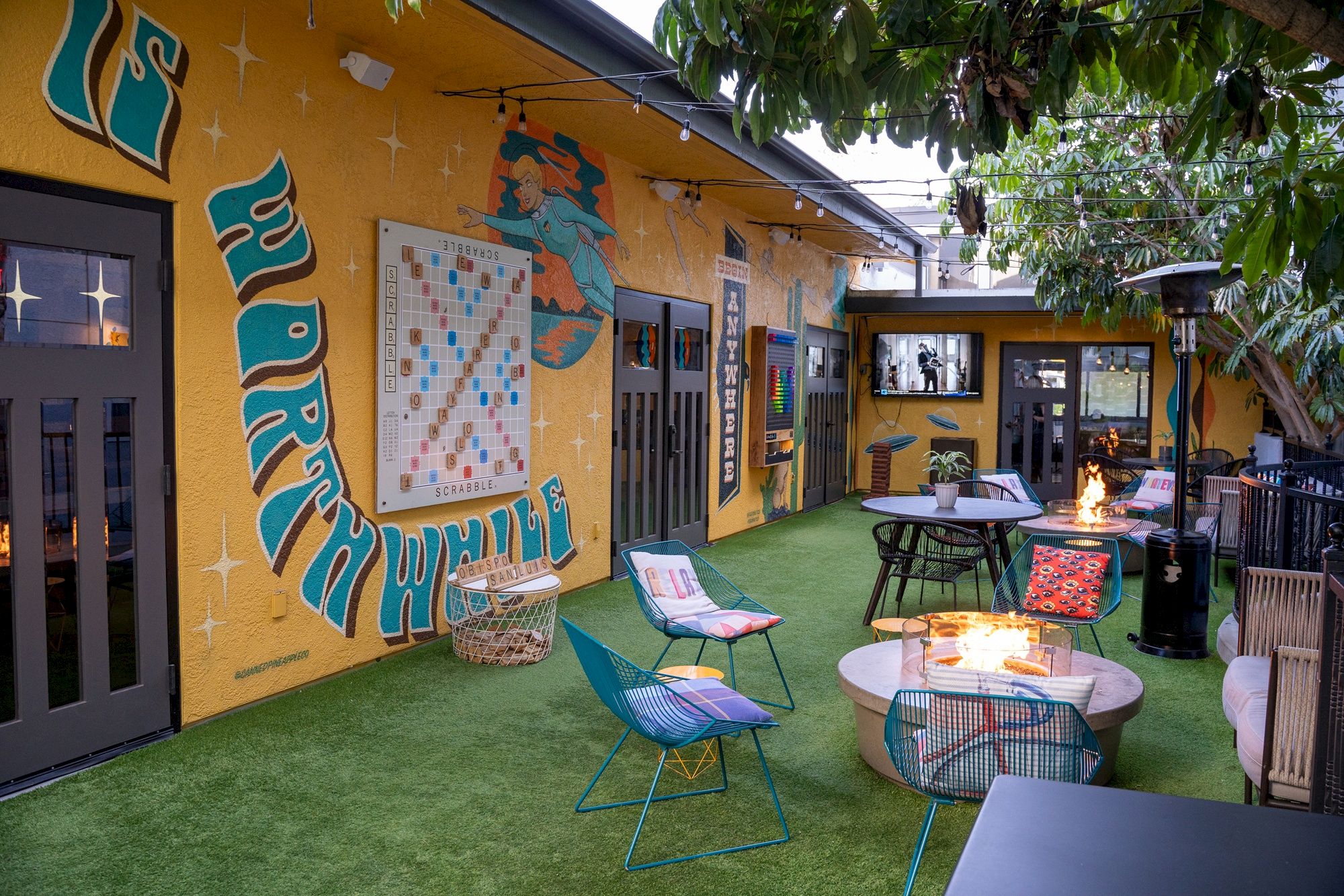 Sunlit patio courtyard with a colorful graffiti mural, a Scrabble board, string lights, lounge chairs, and a cozy firepit.