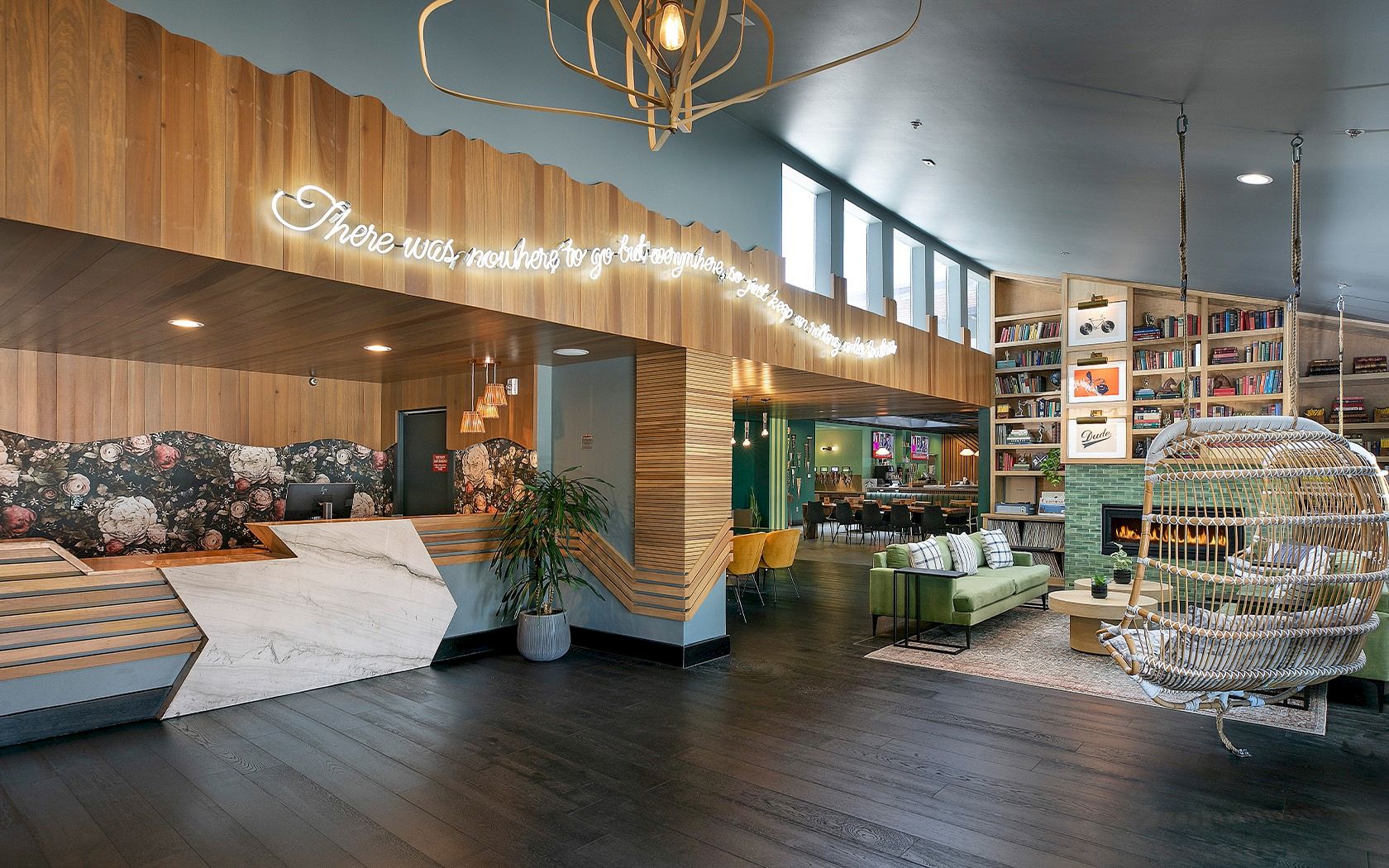A stylish reception area with a wooden desk, warm lighting, a green sofa section, a shelving wall, and a hanging chair in a bright, modern lobby.