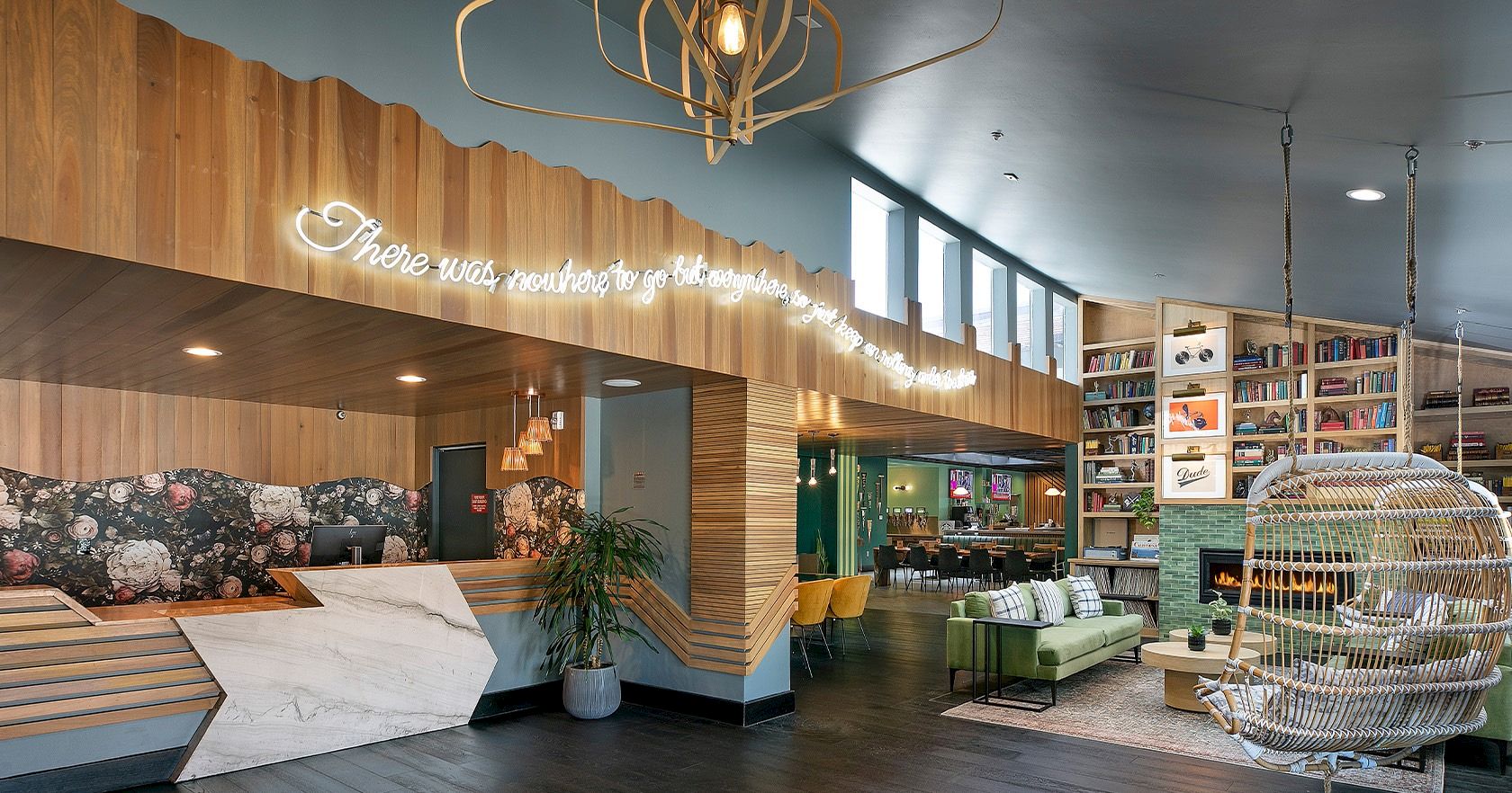 A stylish reception area with a wooden desk, warm lighting, a green sofa section, a shelving wall, and a hanging chair in a bright, modern lobby.