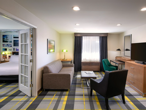 A modern hotel suite with a living area and adjoining bedroom, featuring a gray sofa, green chair, desk, and a flat-screen TV.