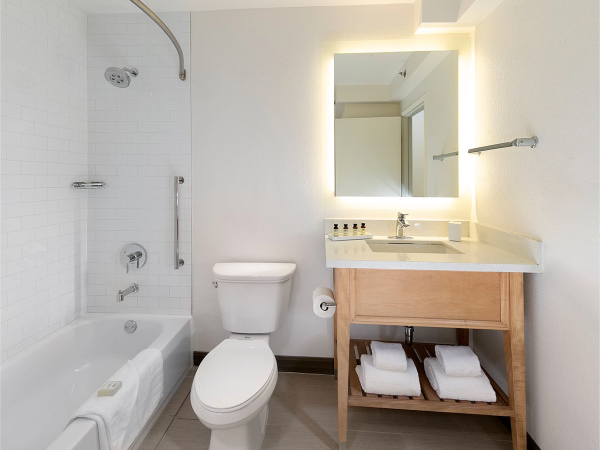 A modern bathroom with a bathtub/shower combo, toilet, and a wooden vanity featuring a backlit mirror and a stacked towel shelf beneath.
