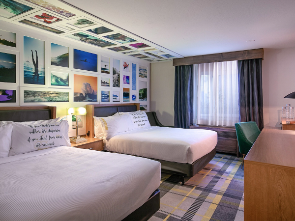 A hotel room with two beds, wall collage of photos above, a desk, TV, window with curtains, and a chair by the desk.