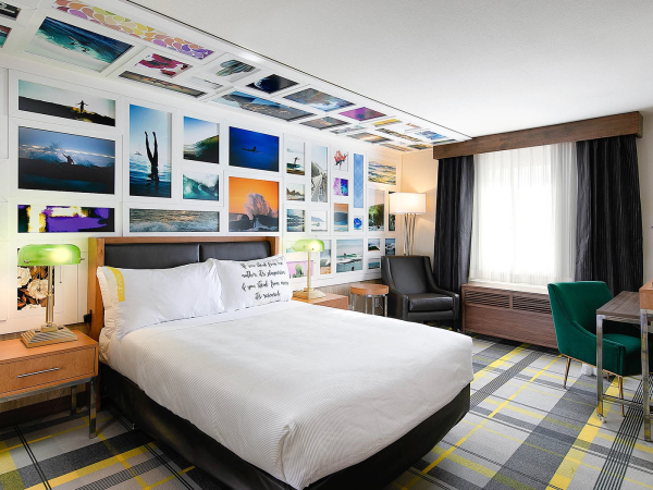 A stylish hotel room with a photo gallery wall, comfortable bed, two nightstands, a desk area, and a mix of green and yellow accents.