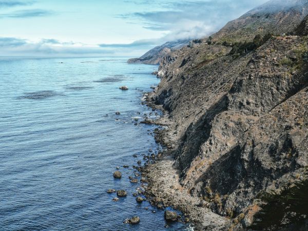 A rocky coastline with steep cliffs meeting calm blue sea, small rocks along the shore, and a clear sky above&mdash;nature's rugged seascape.