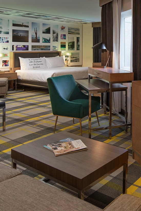 A modern hotel suite with a seating area, coffee table, TV on a wooden dresser, a desk with chair, a green accent chair, and a bed at the back with wall art and photos, all in warm lighting.