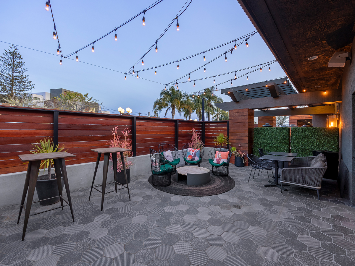 A stylish rooftop patio with hexagonal tile flooring, string lights, tall plant stands, a circular outdoor rug seating area, colorful cushions, and a surrounding privacy fence.