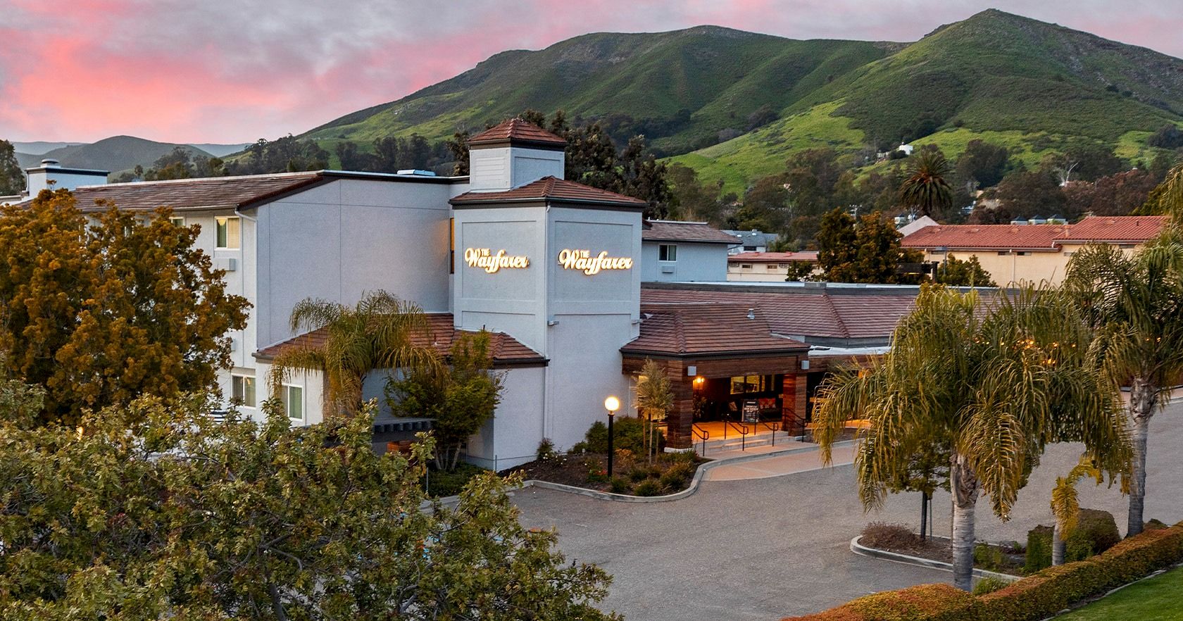 A hillside hotel with a light-blue facade and a sign reading &ldquo;Mauna Loa&rdquo; or similar, surrounded by trees and a parking area at sunset.
