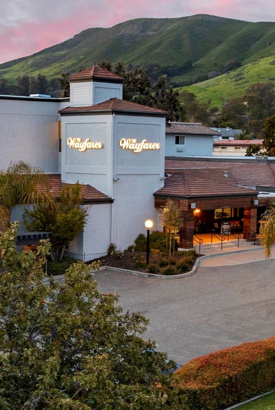 A mid-size hotel or resort building with a sign reading &ldquo;Wine &hellip;&rdquo; (likely a winery-themed inn), surrounded by trees and hills at sunset, hotel entrance lit up.