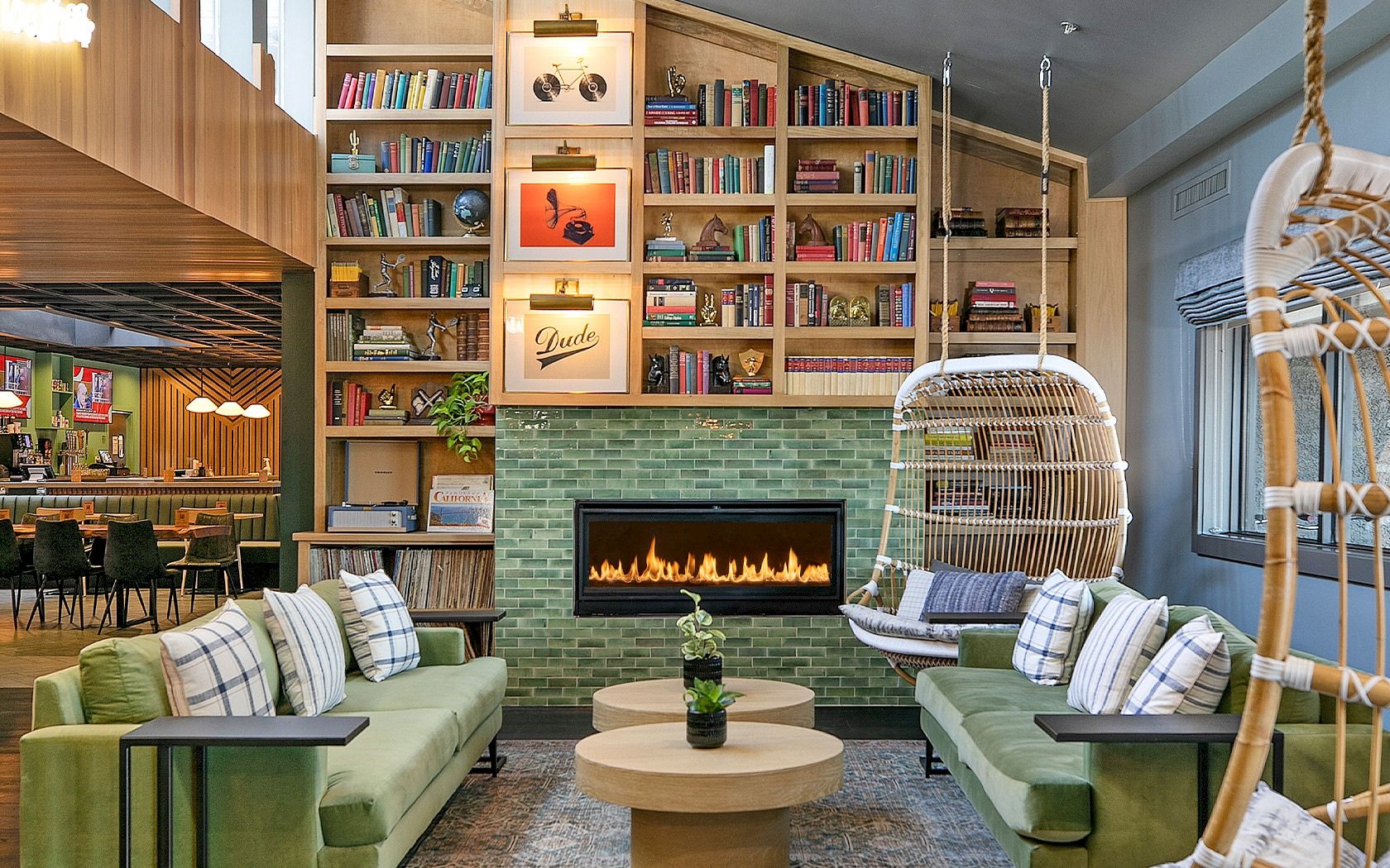 A cozy, modern library-lounge with green sofas, a brick fireplace, hanging chair, and tall book shelves filled with books, creating a warm communal vibe.