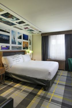 A modern hotel room with a bed, desk, TV, and a wall of colorful framed photos, plus curtains and lamps.
