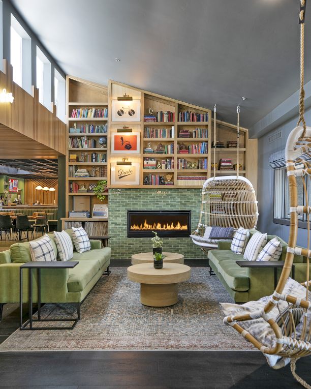 A cozy lounge/library with built-in bookshelves, a lit fireplace, sofas, and a hanging wicker chair by the window.