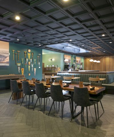 A modern indoor lounge/restaurant with tables, green booths, a bar counter, sports banners, and a lit paneled ceiling.