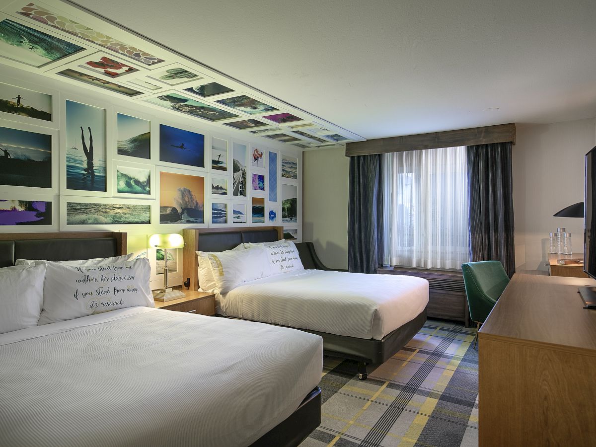 A modern hotel room with two beds, colorful ocean-themed wall art, soft curtains, and a desk by the window.