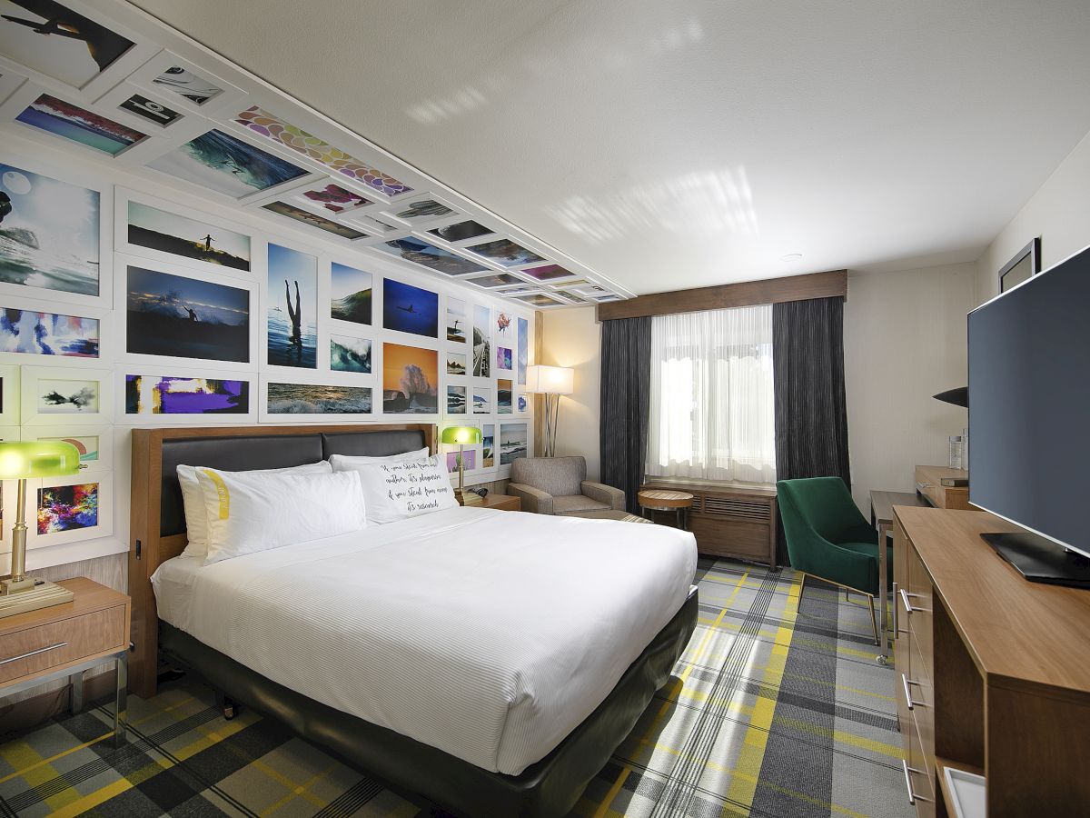 A modern hotel bedroom with a large bed, colorful wall art gallery, green accents, curtains, and a desk/TV setup.