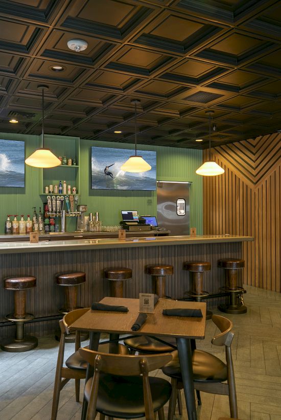 An indoor bar/lounge with a long counter, stools, hanging pendant lights, and two screens showing surfers in the ocean.