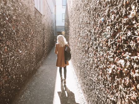 A woman strolls down a narrow alley, holding a bag, as bright sun bounces off walls covered in tiny flyers or glitter.
