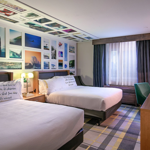 A bright hotel room with two large beds, a collage of travel photos on the wall, a desk, a TV, and a window with blue curtains.