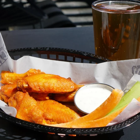 Basket of buffalo wings with dipping sauce and melon slices, plus a glass of beer on a sunny outdoor table.