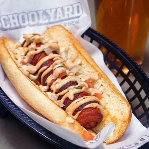 Hot dog loaded with bacon, onions, and sauce in a toasted bun, served in a basket with a drink in the background.