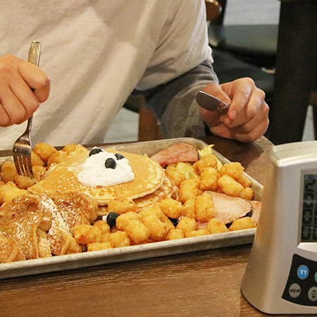 A person eats a pancake plate with a large pancake shaped like a bear face, hash browns, bacon, syrup, and a digital timer nearby.