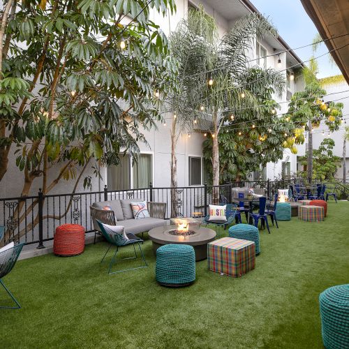 A stylish outdoor lounge area with grassy ground, colorful poufs, low tables, string lights, and multiple seating nooks next to a building.