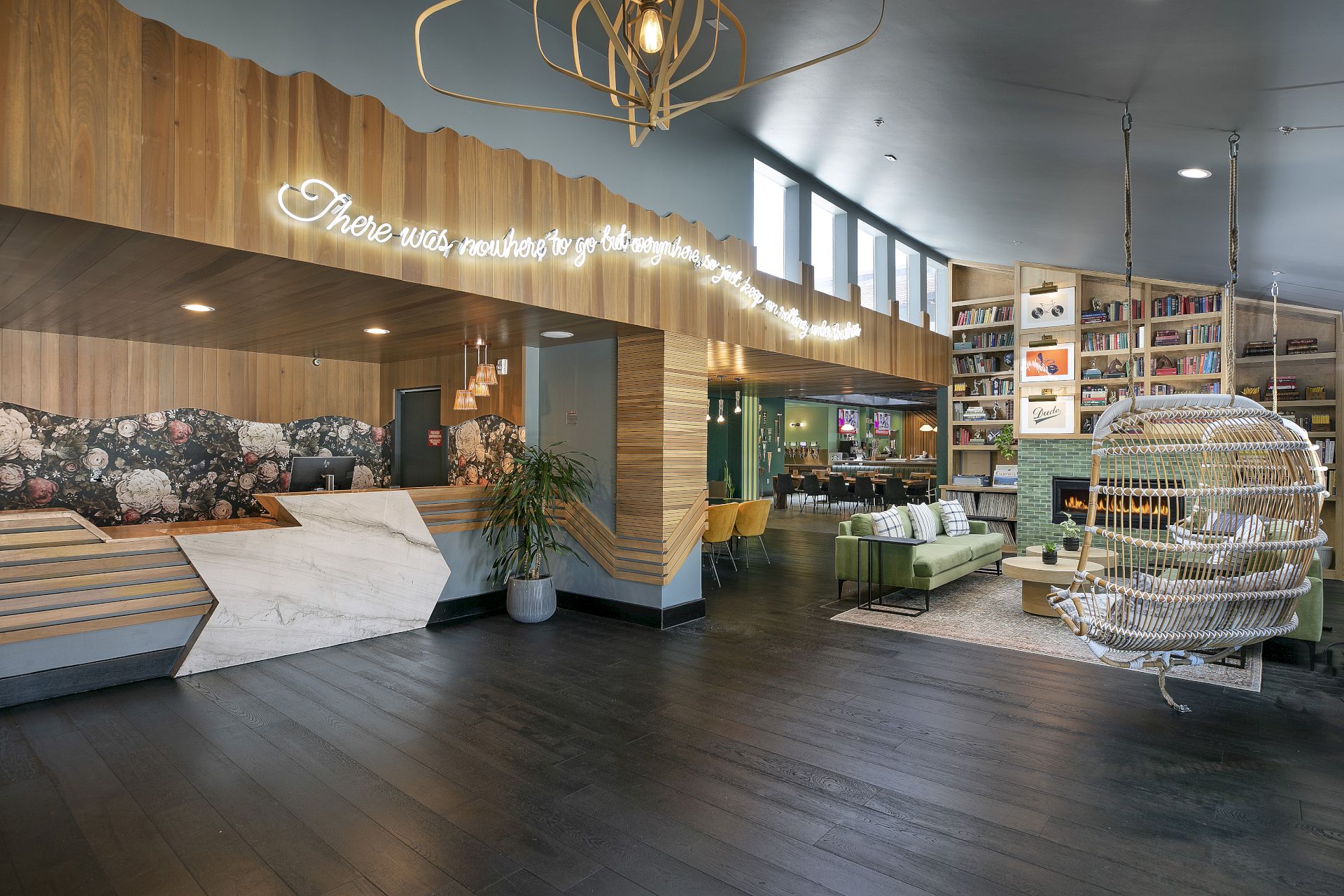 A cozy modern caf&eacute; lounge with bookshelves, a fireplace, rattan chairs, floral wall art, and hanging lights overhead.