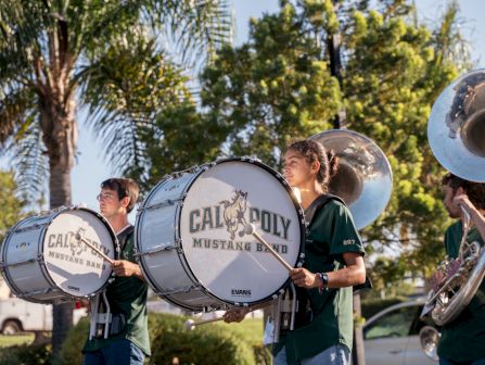 A marching band plays outdoors with drummers holding large bass drums labeled &ldquo;Cali Poly Mustang Band,&rdquo; alongside brass instruments.